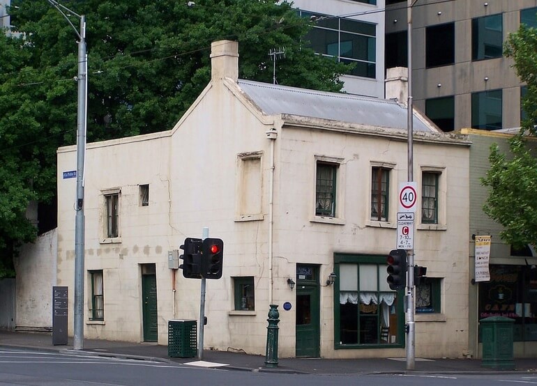 Small render-clad heritage corner building standing beside a modern office tower